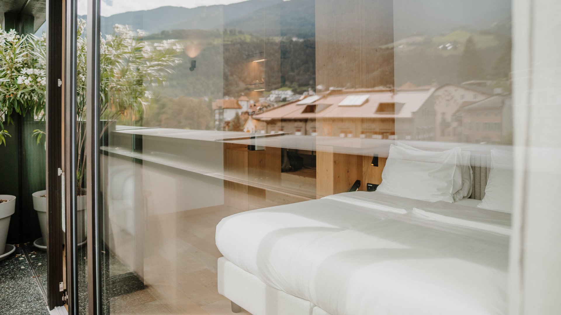 Blick durch Glas auf ein helles Bett in einem Zimmer mit Bergblick