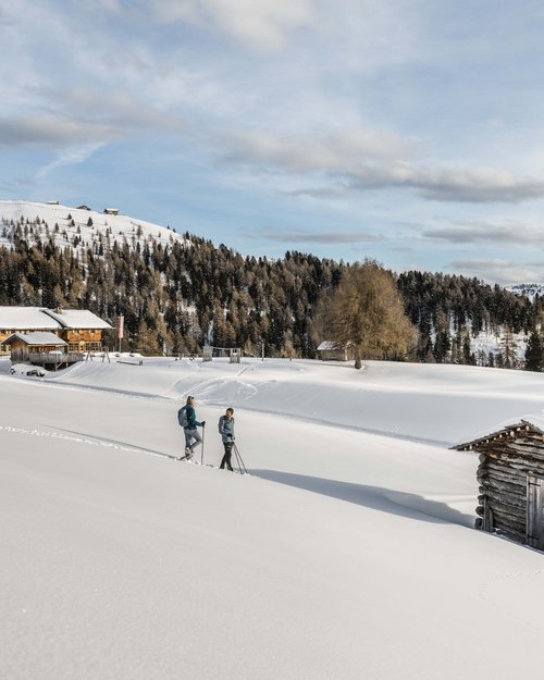 Two people snowshoeing in a snowy mountain landscape with cabins