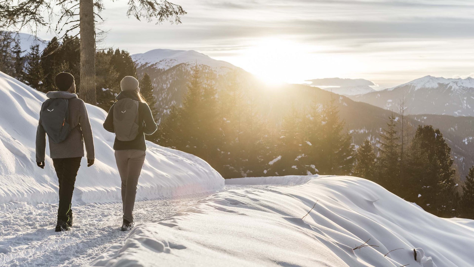 Two hikers walking on snowy trail at sunset in the mountains